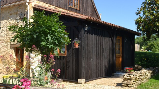A rustic wooden house with a stone foundation, a wooden door, and a small garden with pink flowers. - L'écrin du Val de Louyre, in the heart of Périgord Noir, with private pool (Val de Louyre et Caudeau)