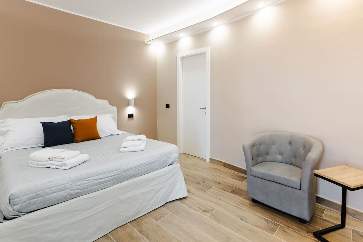 A neatly made bed with pillows, a chair, and a small table in a room with wooden flooring and a white door.
