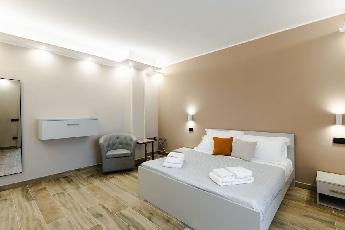 A modern hotel room with a large bed, a chair, a desk, and a mirror on the wall.