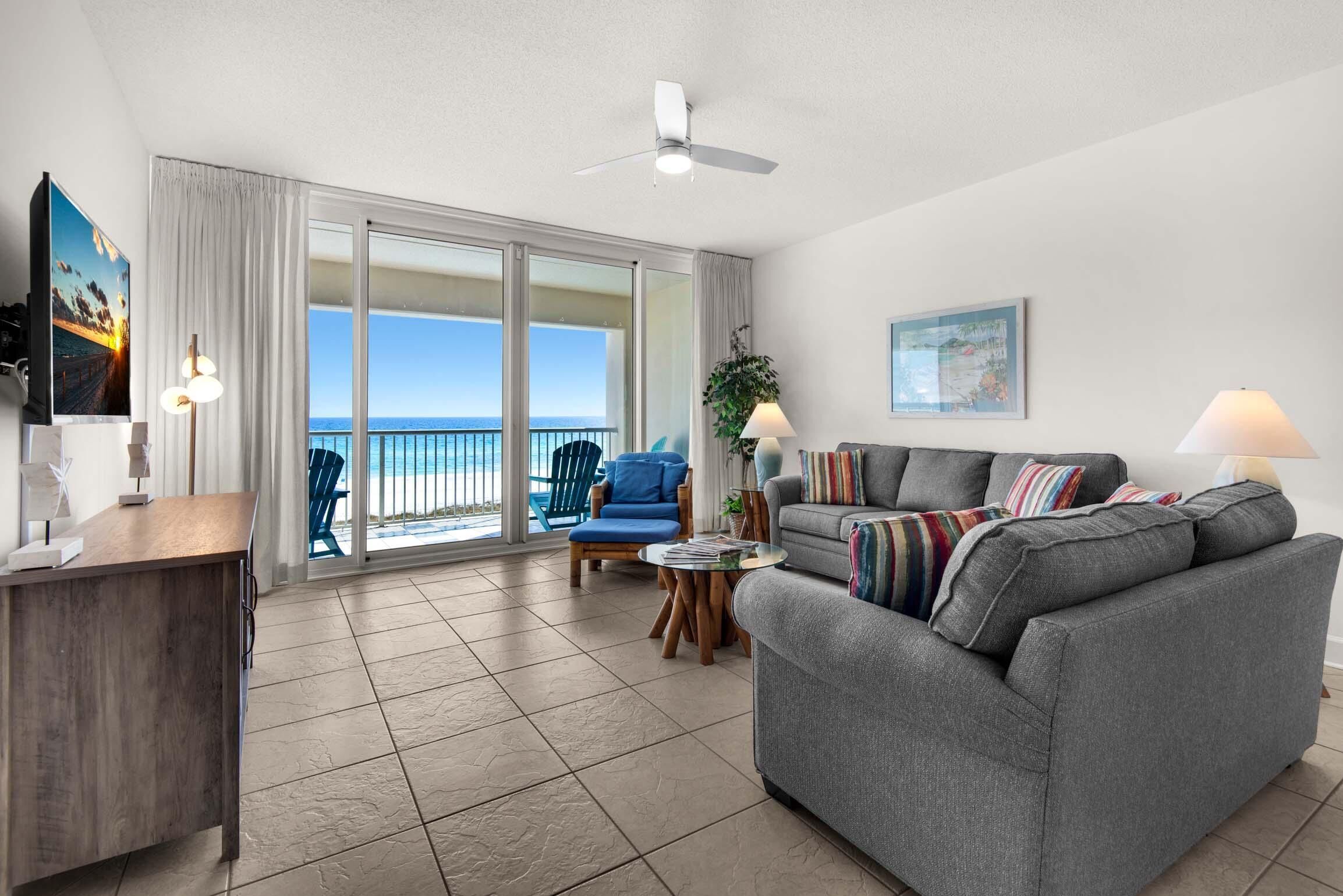 A spacious living area with a gray sofa, a blue chair, a wooden desk, and a large sliding glass door leading to a balcony with a view of the ocean.