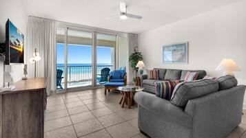 A spacious living area with a gray sofa, a blue chair, a wooden desk, and a large sliding glass door leading to a balcony with a view of the ocean.