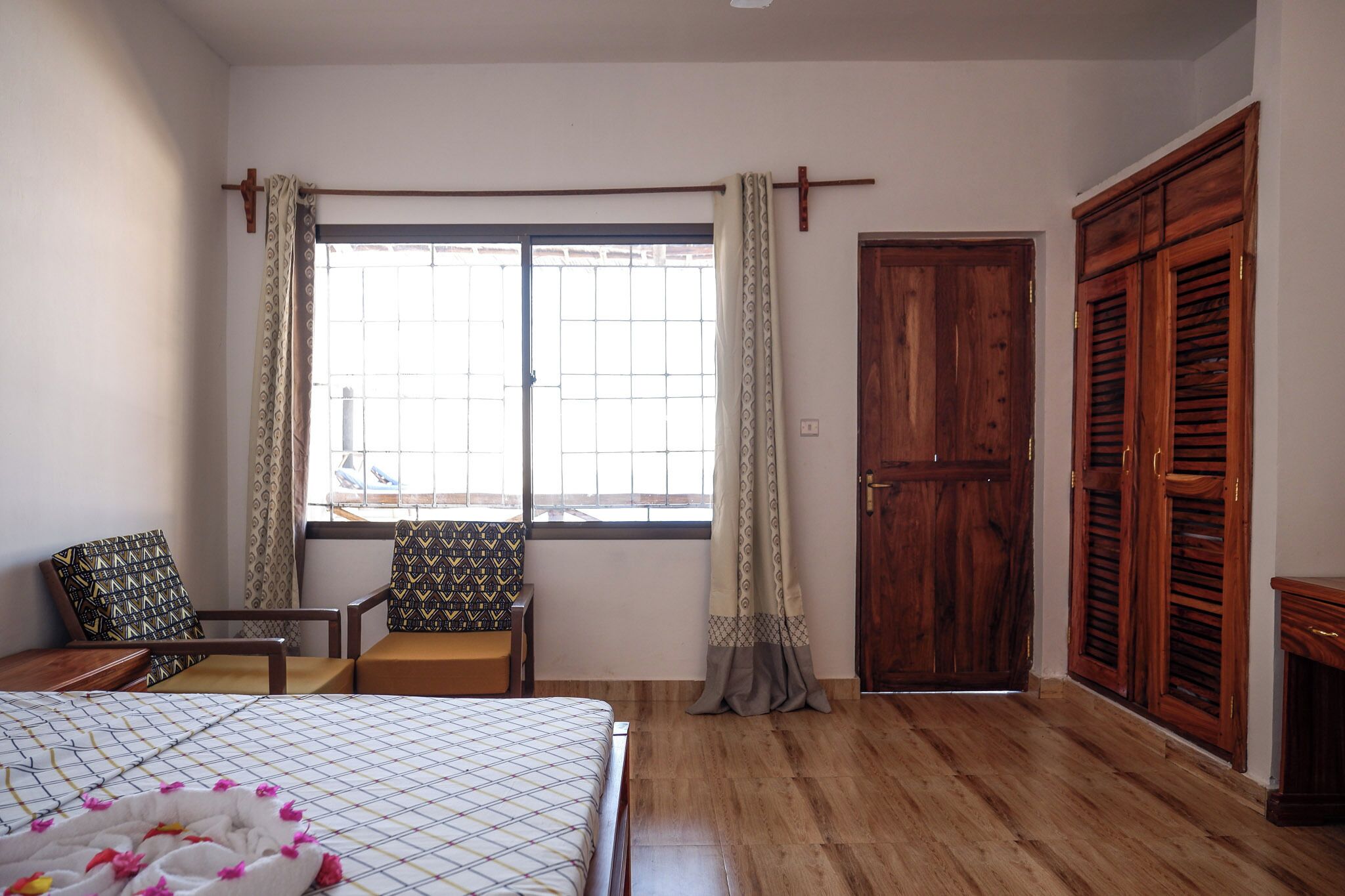 A bedroom with a bed, two chairs, a wooden wardrobe, and a window with curtains.
