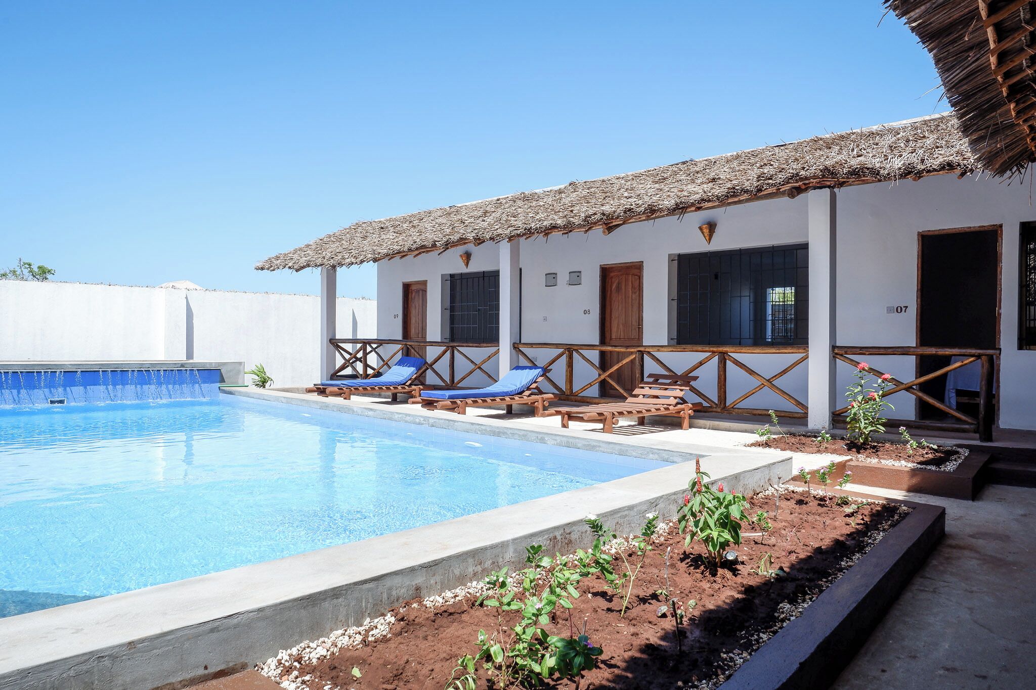 Un área de piscina con un edificio de techo de paja, sillones de descanso y un pequeño jardín.