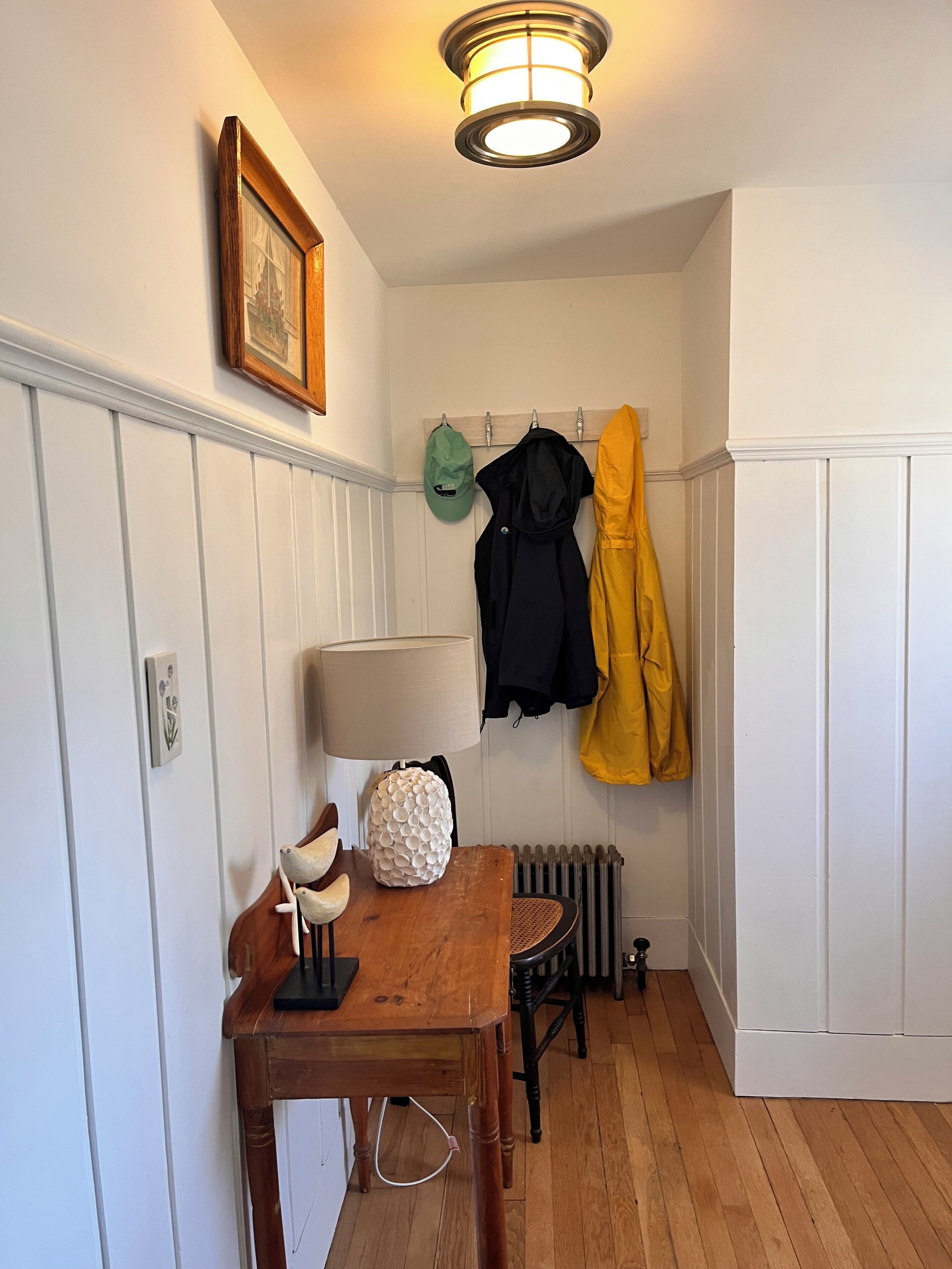 A corner room with a wooden table, a lamp, a chair, a radiator, and a coat rack with jackets.