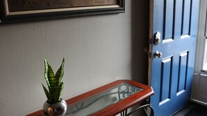 Un pasillo con una puerta azul, un cuadro enmarcado y una mesa con superficie de vidrio y una planta.