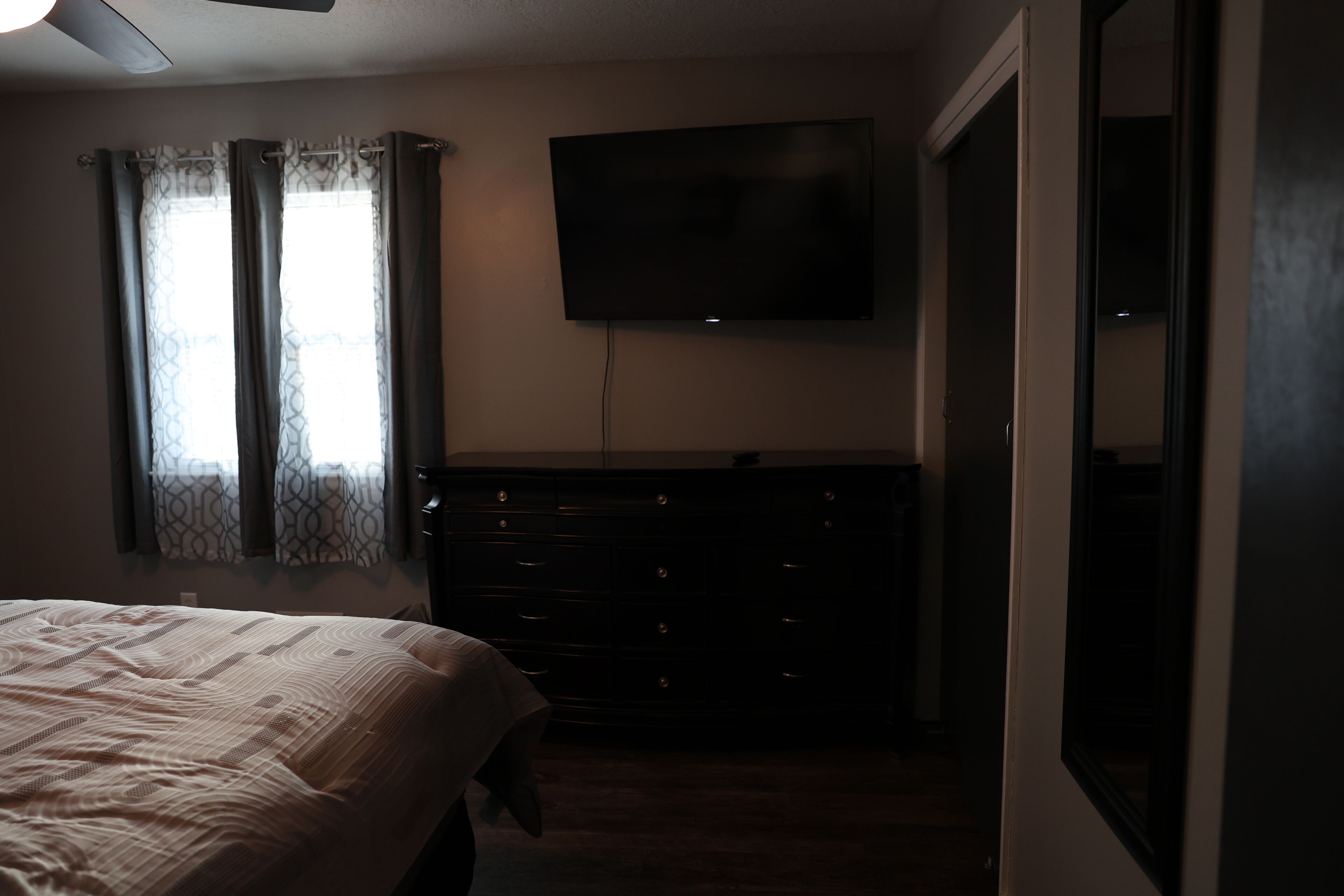 Ein Schlafzimmer mit Bett, Kommode, Fernseher und einem Fenster mit Vorhängen.