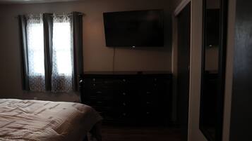 Ein Schlafzimmer mit Bett, Kommode, Fernseher und einem Fenster mit Vorhängen.