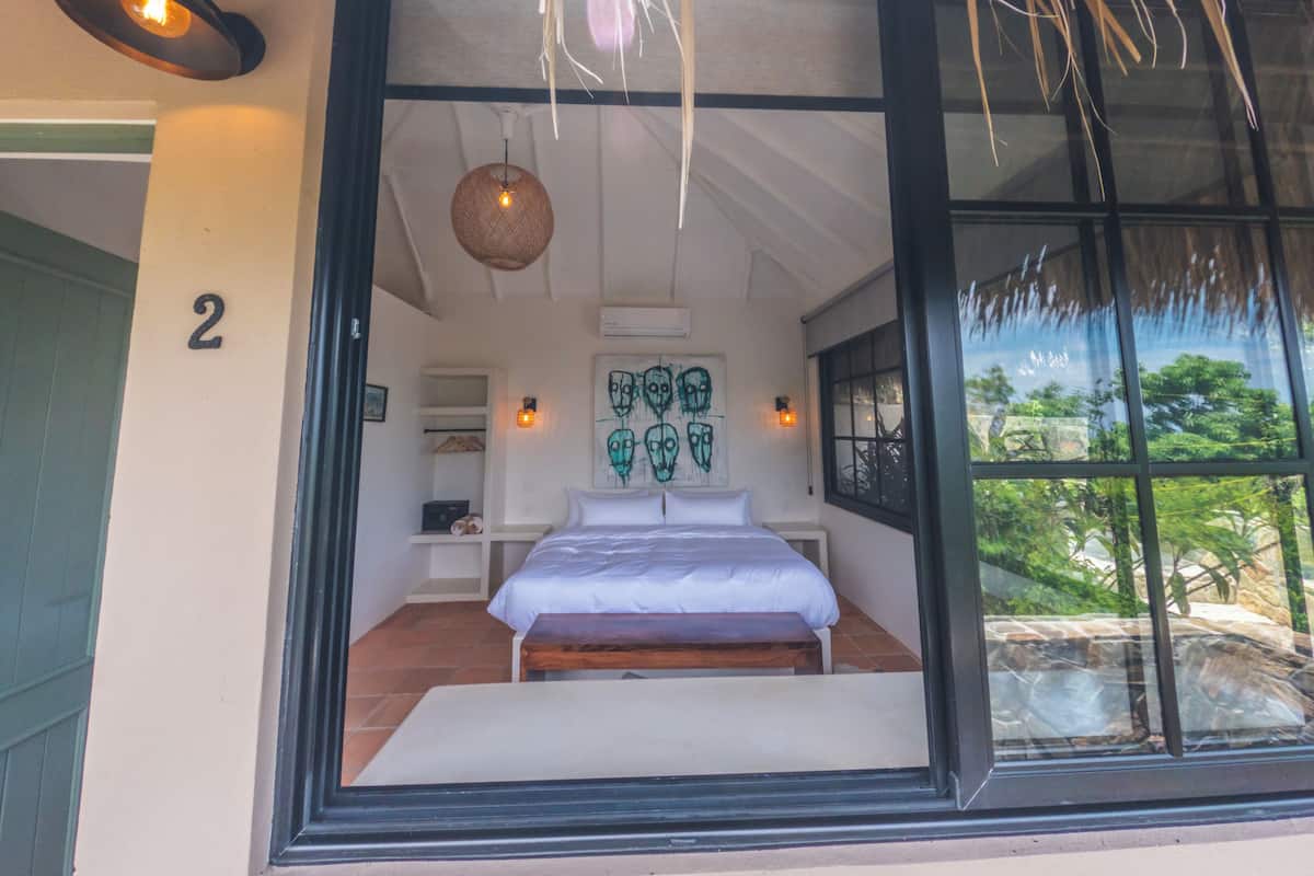Ein Schlafzimmer mit einem Bett, einem Wandregal, einer Hängeleuchte und einem großen Fenster mit Blick auf Grün.