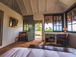 Una habitación con cama, escritorio, silla, un cuadro y ventana con vista a follaje.