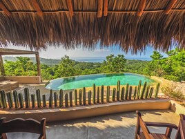 Una piscina con cubierta de madera y plantas de cactus, rodeada de abundante vegetación y vistas al océano.