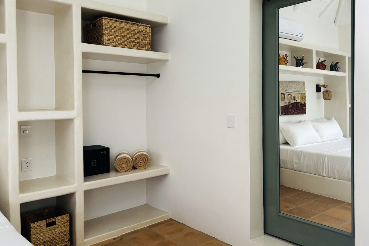 Quarto com parede branca, cama com roupa de cama branca, uma pequena prateleira com objetos decorativos e um espelho refletindo uma cama e um ar condicionado fixado na parede.