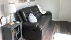 A dark leather sofa with a white throw blanket and pillows, a small side table with a lamp and decorative items, and two nautical-themed paintings on the wall. - Charming shipping container home with lake view and boat ramp. (Cookson)