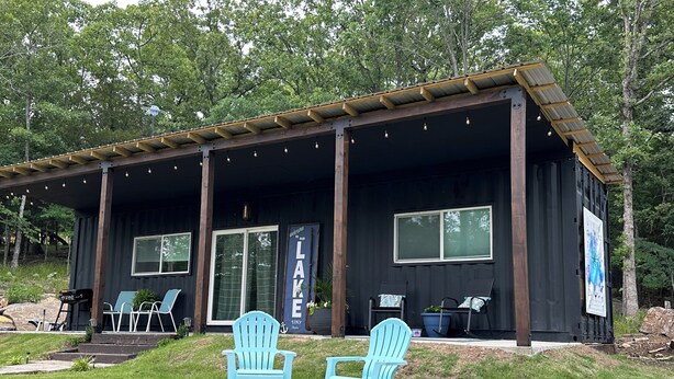 A cabin with a covered porch, two Adirondack chairs, a fire pit, and a welcome sign. - Charming shipping container home with lake view and boat ramp. (Cookson)