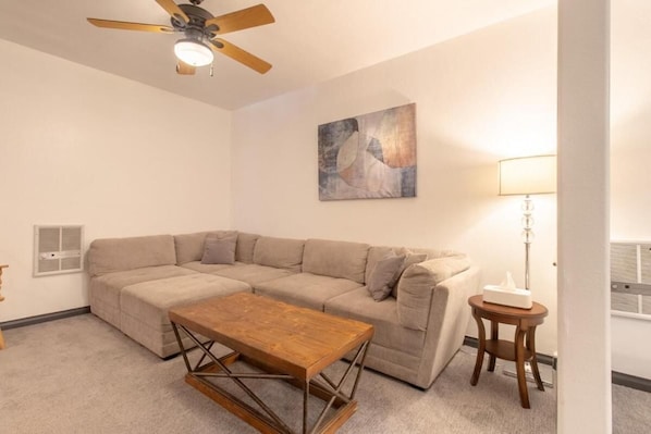 A living room with a beige sectional sofa, a wooden coffee table, a ceiling fan, a wall-mounted lamp, and a painting.