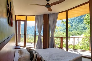 A bedroom with a bed, curtains, a ceiling fan, and large windows offering a view of a mountainous landscape. - Hotel Anukwe (Santa Marta)
