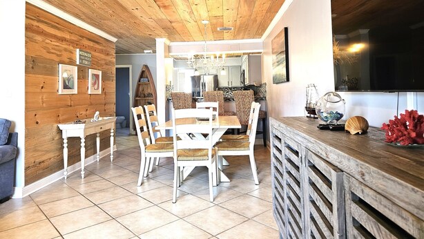 A modern kitchen with wooden cabinets, a dining area with white chairs, and a TV mounted on the wall. - 11 Drift - 2BR $700+ in freebies incl golf, pool (Miramar Beach)