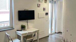 A room with a white desk, a chair, a flat-screen TV, a window with blinds, and a door with a glass panel. - Macallè House Letojanni - Taormina (Letojanni)
