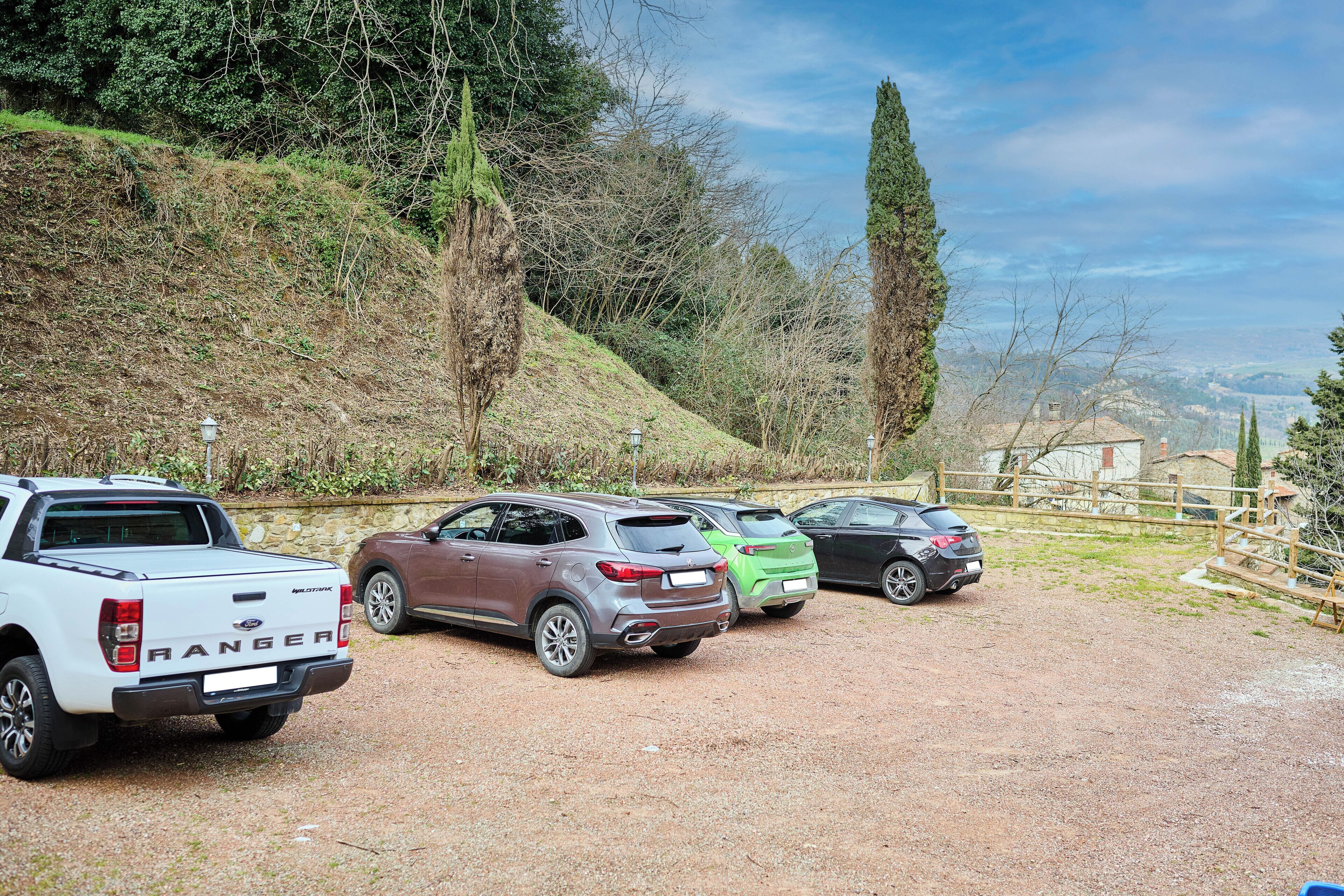 Ein Kiesparkplatz mit mehreren Fahrzeugen, darunter ein weißer Pritschenwagen und ein brauner SUV, auf einem Hügel gelegen, mit Blick auf ein Haus und Bäume in der Ferne.