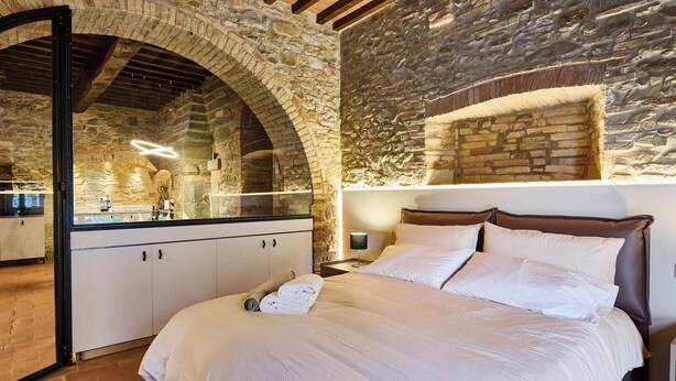 A bedroom with a stone wall, a wooden ceiling, a bed with white linens, and a nightstand with a lamp. - The Crusher (whirlpool) (Rivalto)