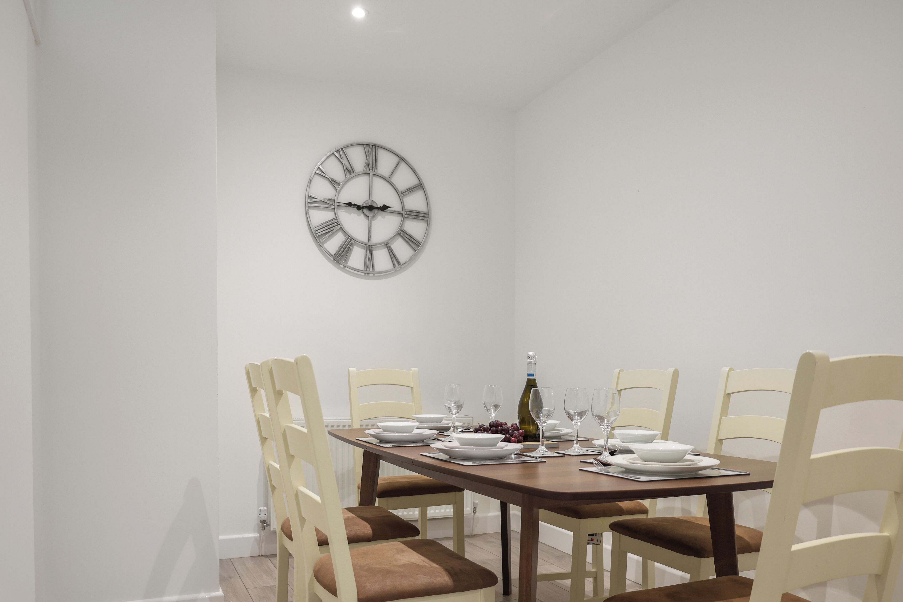 A dining room with a wooden table set for four, white chairs, a wall clock, and a bottle of wine.