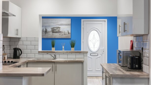 A modern kitchen with white cabinets, a blue accent wall, a painting of a boat, and a door with a circular window.