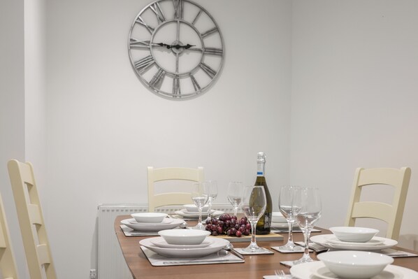 A dining table set for six with white plates, bowls, and glasses, a bottle of wine, and a clock on the wall. - Stunning 5 Bed property in Newbiggin -by-the-sea (Newbiggin)