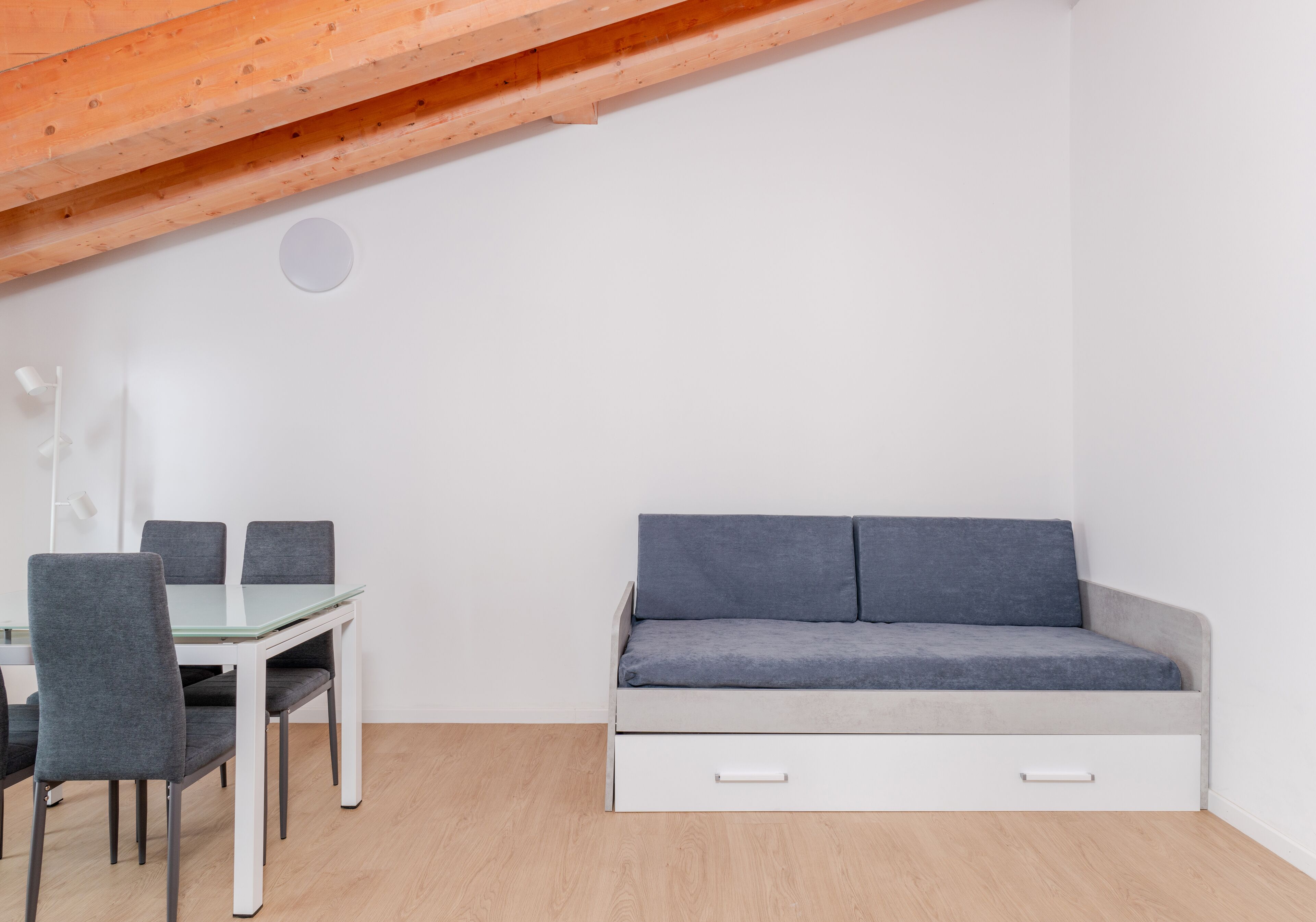 Una habitación con una mesa de comedor de cristal, cuatro sillas grises, un sofá gris con cajones y una viga de techo de madera.