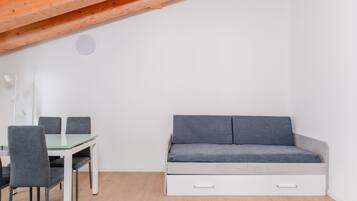 Una habitación con una mesa de comedor de cristal, cuatro sillas grises, un sofá gris con cajones y una viga de techo de madera.