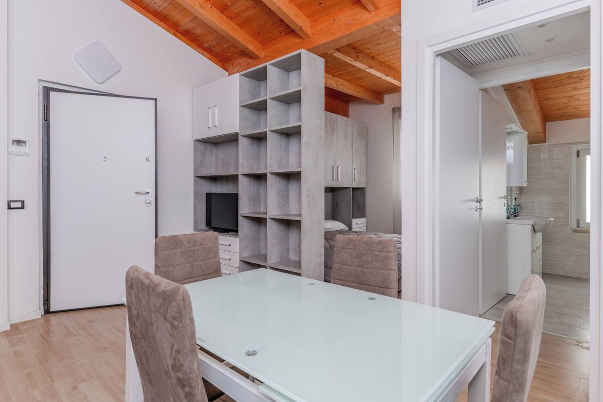 A modern room with a glass table, cushioned chairs, a built-in shelving unit, and a kitchen area visible through an open door.