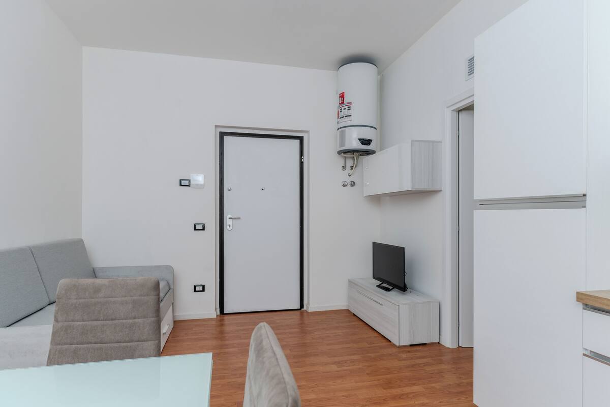 A modern, minimalistic room with a grey sofa, a flat-screen TV, a white cabinet, and a water heater mounted on the wall.