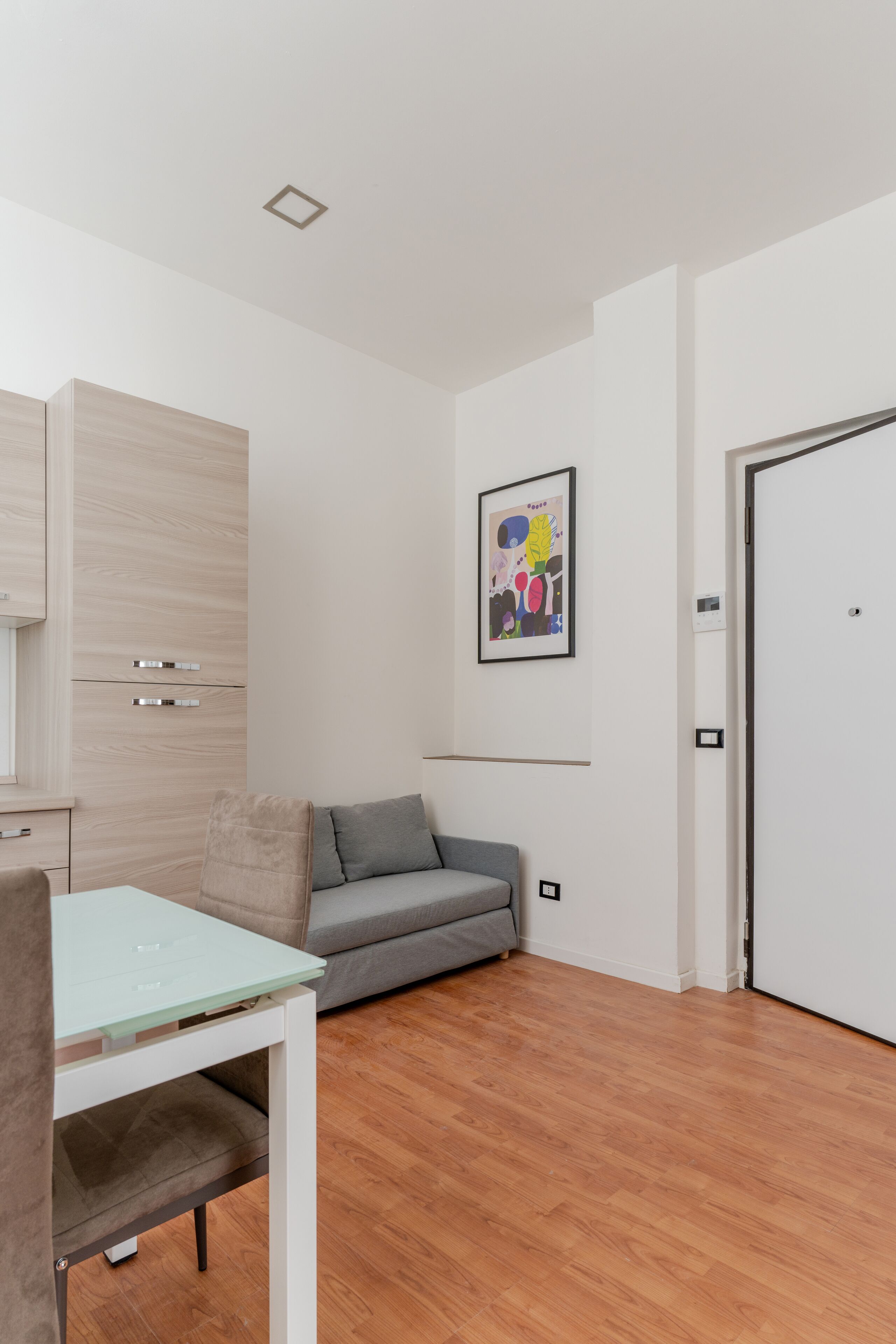 Una habitación moderna con piso de madera, una mesa de vidrio, un sofá gris y una puerta blanca.