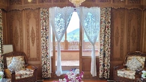 An opulent room with a large chandelier, ornate wooden paneling, and a view through a window with floral curtains. - New Alzira Heritage Group of Houseboats  (Srinagar)
