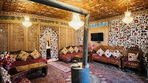 A traditional room with intricate wooden carvings, a central wood-burning stove, and multiple seating areas with patterned cushions. - New Alzira Heritage Group of Houseboats  (Srinagar)