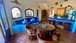Un área de estar acogedora con un sofá azul, una mesa redonda de vidrio, sillas de mimbre y una cocina al fondo.