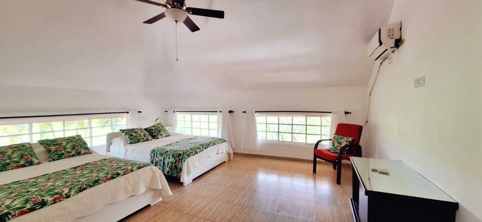 Una habitación con dos camas, un ventilador de techo, una silla roja y un escritorio.