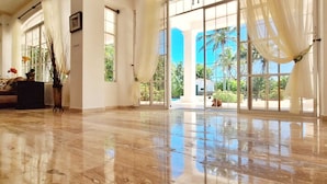 A spacious room with a polished marble floor, large windows with sheer curtains, and a view of palm trees and a clear blue sky. - 9th Hole Villa (Guayacanes)