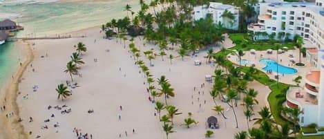 Un complejo turístico en la playa con una extensa playa de arena, palmeras y una zona de piscina.