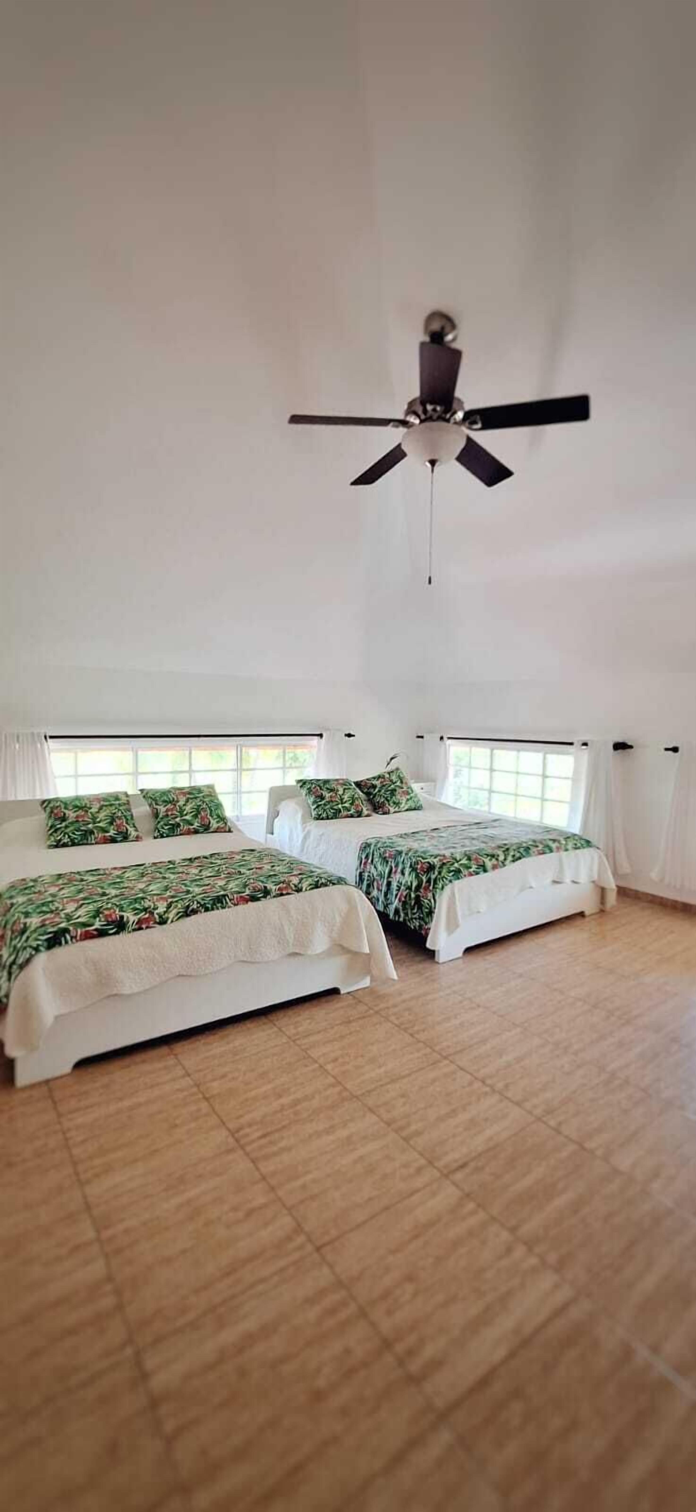 Un dormitorio amplio con dos camas, ventilador de techo y ventanas grandes.