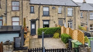 A residential building with multiple windows, a front door, and a small garden area with a wooden fence and some plants. - Cosy Walkers Retreat in Meltham Doggie Friendly (Holmfirth)