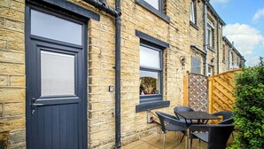 A brick house with a dark blue door and windows, a patio with a table and chairs, and a wooden fence. - Cosy Walkers Retreat in Meltham Doggie Friendly (Holmfirth)