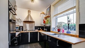 A modern kitchen with black cabinets, a stainless steel range hood, a large window with blinds, and a view of greenery outside. - Cosy Walkers Retreat in Meltham Doggie Friendly (Holmfirth)