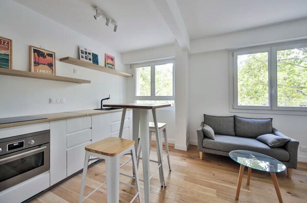 A modern kitchen with white cabinets, a built-in oven, and a wooden countertop. There is a dining table with a chair, a gray sofa, and a glass coffee table. Large windows provide natural light and a view of trees.