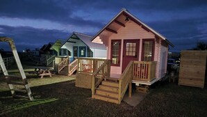 Een roze blokhut met een houten trap en terras, omgeven door een grasveld met een picknicktafel en een houten constructie.