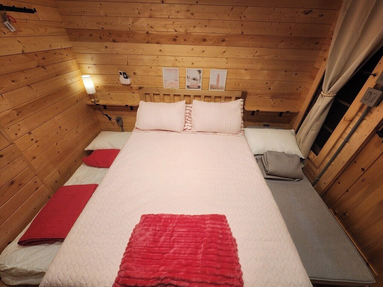 A wooden cabin room with a bed, a red blanket, and a small table with a lamp.