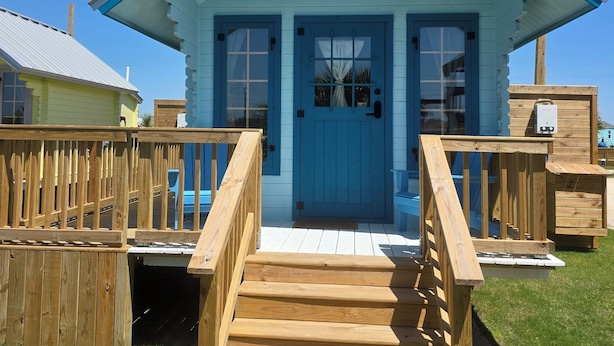 Un petit bâtiment en bois, avec une porte bleue et un escalier en bois menant à l’entrée.