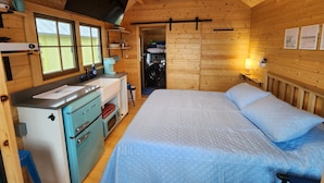 Une petite pièce de cabane en bois comprenant un lit, un coin cuisine, un lave-linge et une fenêtre.