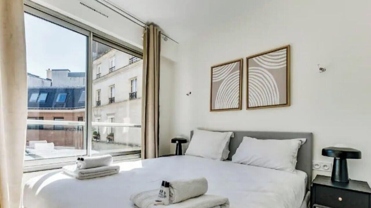 Ein Hotelzimmer mit großem Fenster, einem Bett mit weißen Leinen, zwei an der Wand befestigten Kunstwerken und einem Nachttisch mit Lampe.