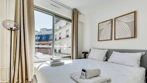 Habitación de hotel con una cama grande, una mesita de noche, una lámpara de pie y vistas de la ciudad a través de un ventanal amplio.