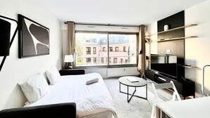 Habitación de hotel con cama, escritorio, silla, mesita, televisión y ventana con vistas a edificios.