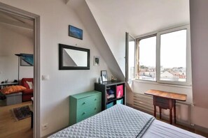 A bedroom with a bed, a desk, a window with a city view, a green dresser, and a mirror on the wall.